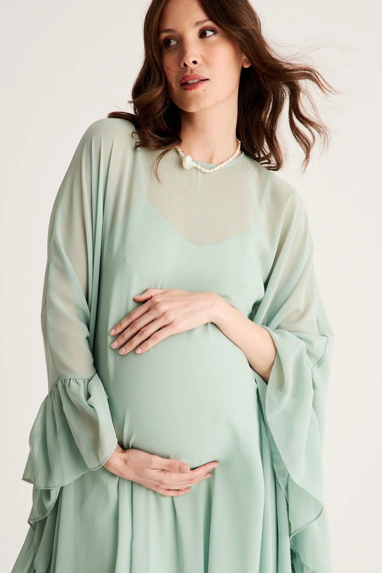 Mujer embarazada con vestido fluido verde agua y mangas con volantes, sosteniéndose la barriga con ambas manos, luciendo un collar de perlas.